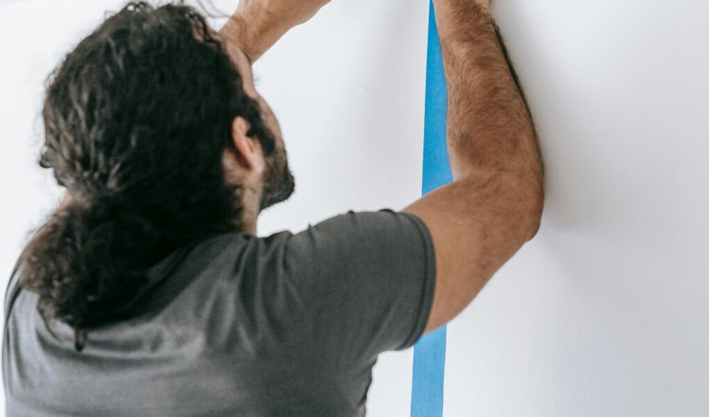 A man applying blue tape on a wall during a home renovation project, showcasing painting preparation.