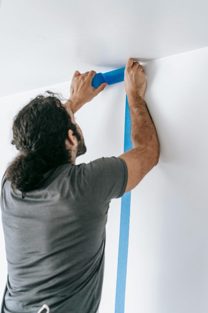 A man applying blue tape on a wall during a home renovation project, showcasing painting preparation.